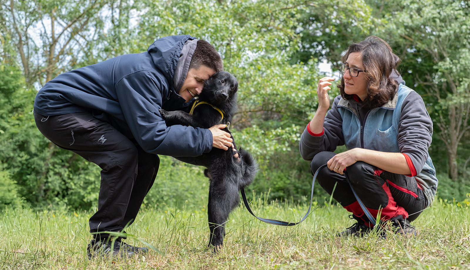 Mariona y Elena abrazando a su perro Benito en un campo de entrenamiento en el mes de septiembre, con césped y árboles verdes.